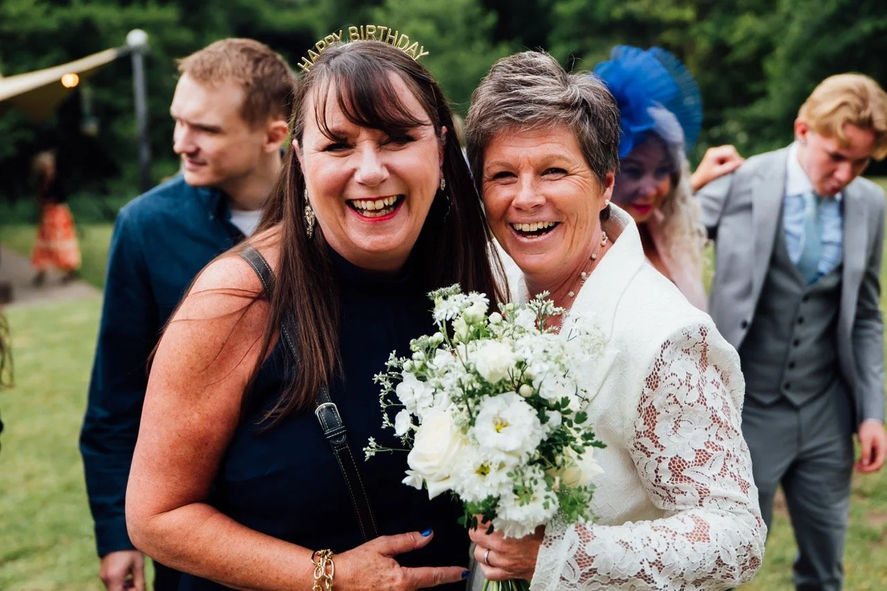 Two women smiling at a celebration, with one holding a bouquet of white flowers; one wears a white lace dress and a 'Happy Birthday' headband, while the other wears a black top; background includes two men and a woman with a blue hat, outdoors with t