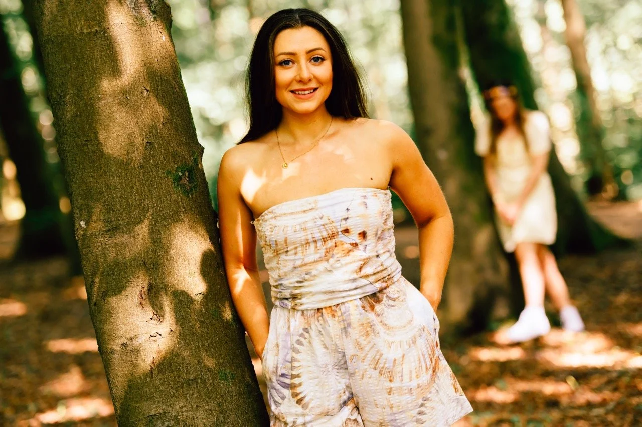A young woman with long dark hair wearing a strapless dress with a brown and white pattern, standing outdoors in a forest, smiling at the camera with other woman blurred in the background.