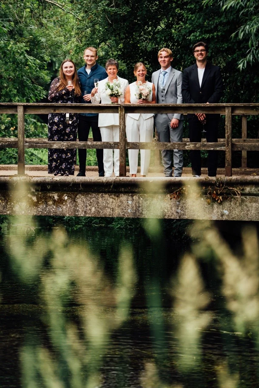 Group of six people standing on a wooden bridge outdoors, celebrating with glasses of champagne and flowers, surrounded by lush greenery.