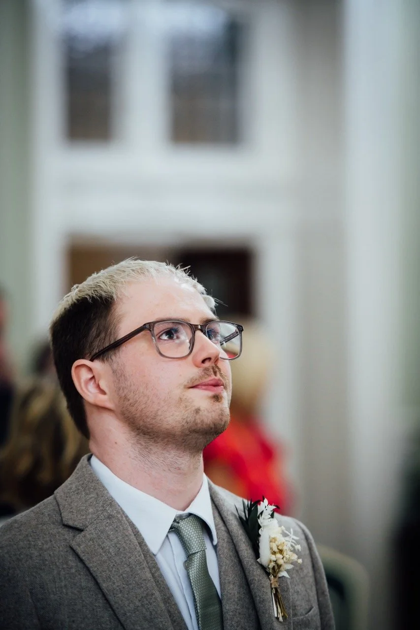 A young man with glasses wearing a gray suit, white shirt, and gray tie, with a boutonniere on his lapel, attending a formal event indoors.