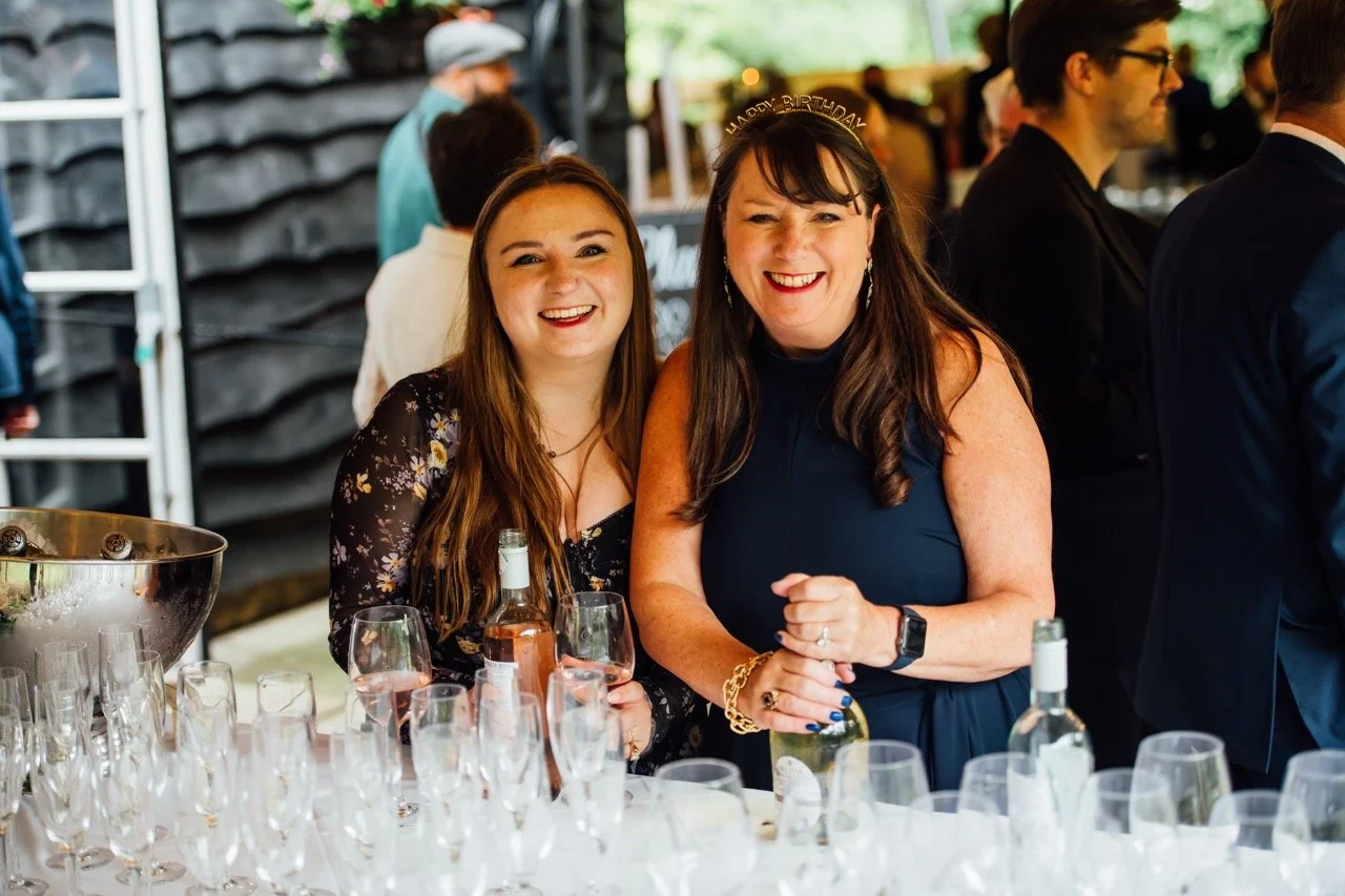 Two women smiling at a celebration, one wearing a 'Happy Birthday' headband, surrounded by glasses and bottles on a table.