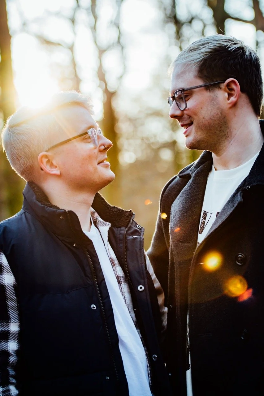 Two men standing close together outdoors in a forest, looking into each other's eyes and smiling, with sunlight filtering through trees in the background.