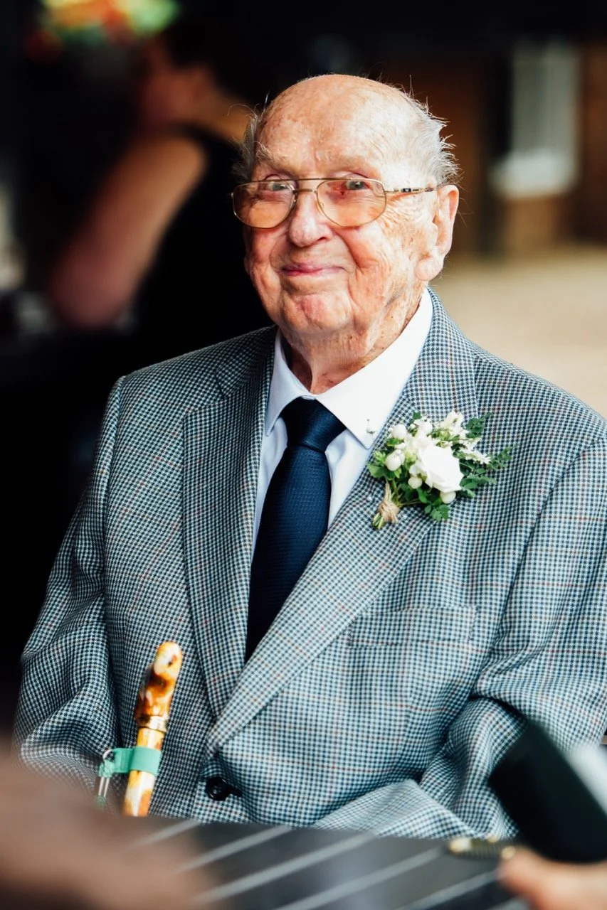 An elderly man with glasses, wearing a checkered suit, white shirt, and dark tie, smiling softly. He has a white boutonniere pinned to his lapel. He is holding a cane with a carved handle, and a blurred background suggests an indoor setting, possibly