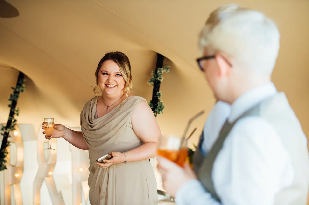 A woman in a beige dress smiling and holding a glass of champagne at a celebration, with a man in glasses and a light-colored shirt in the background holding a drink.
