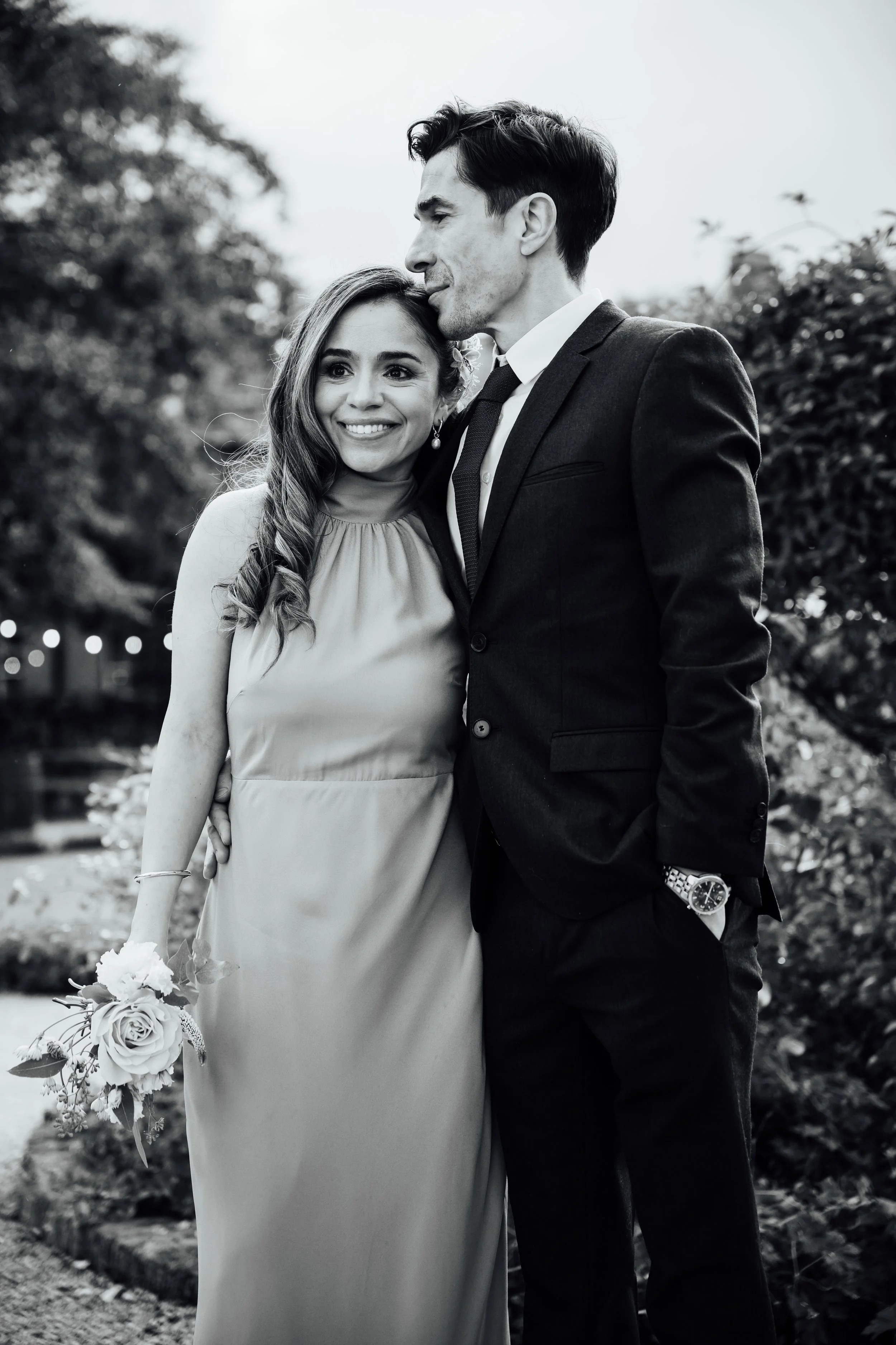 A black-and-white photo of a couple in wedding attire outdoors. The woman is smiling, holding a flower bouquet, and has long, wavy hair. The man is wearing a dark suit, white shirt, and watch, gently kissing the woman's forehead. The background shows