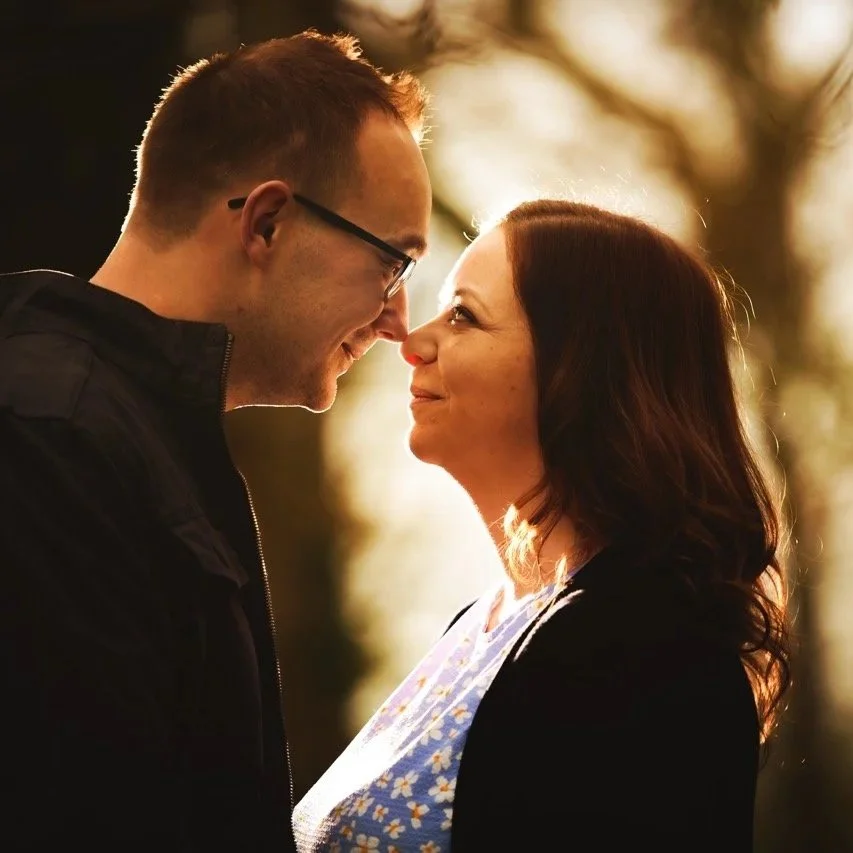 A young man and an older woman face each other tightly outdoors, smiling and touching foreheads, with sunlight and trees in the background.