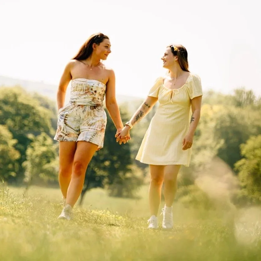 Two women holding hands and smiling while walking in a park on a sunny day.