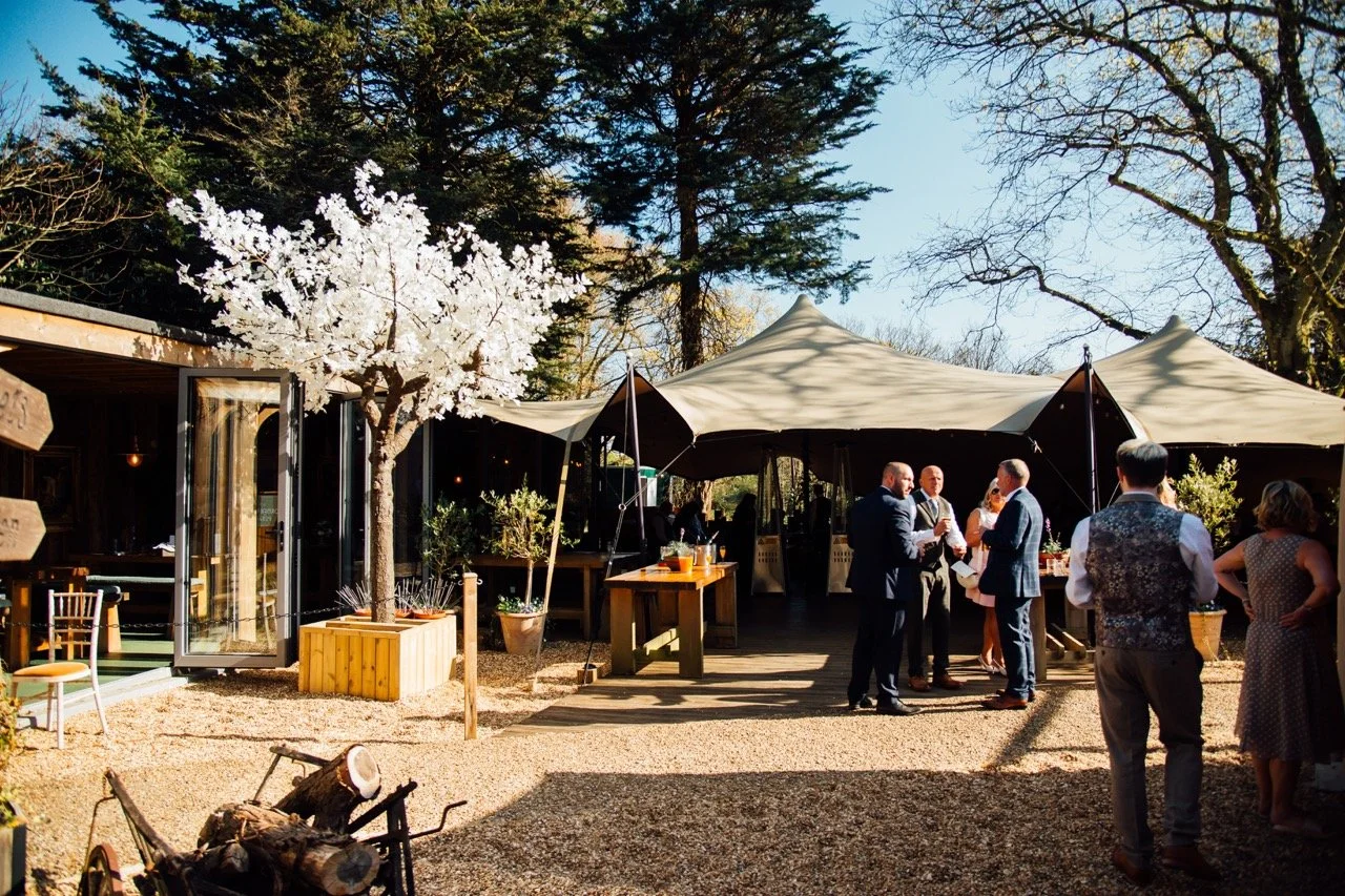 People gather under a large outdoor tent at a social event or wedding, with trees and a building in the background on a sunny day.