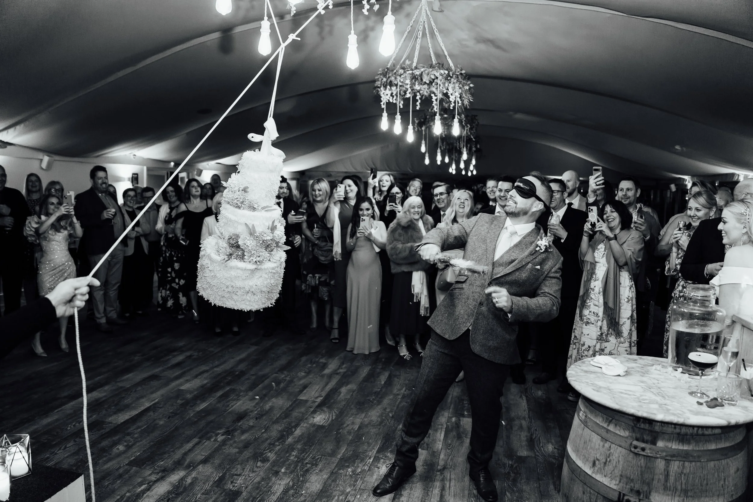 A group of people at a wedding celebration watching a cake smashing event. The groom is blindfolded, holding a plate, ready to smash a cake hanging from a rope. The crowd is smiling and capturing the moment with their phones in an indoor venue with f