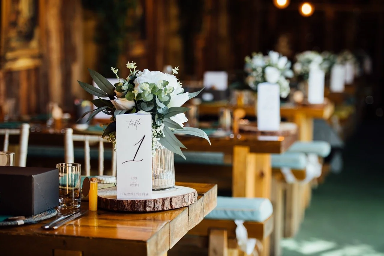 Wedding table centerpiece with white flowers and greenery on a wooden log base, table number card, and place setting in a rustic indoor venue with wooden chairs and warm lighting.