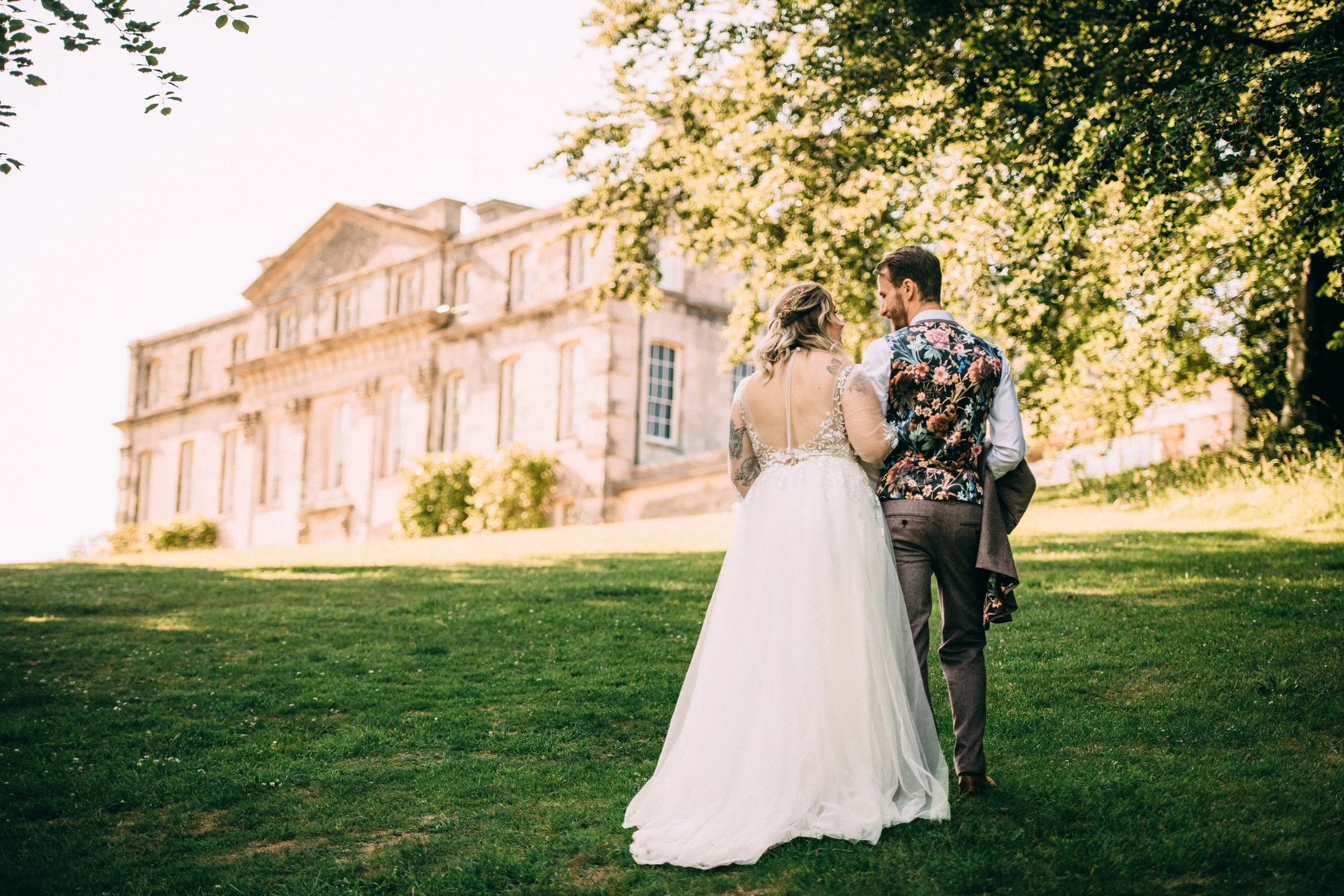 A bride and groom walking on green grass near a large historic building, with the bride wearing a white wedding dress and the groom in a floral vest and brown trousers, under a leafy tree.