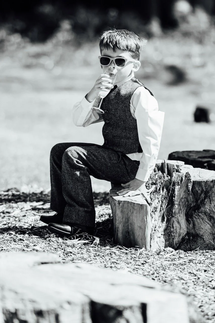A young boy in formal attire, wearing sunglasses, sitting on a tree stump outdoors, drinking from a glass.