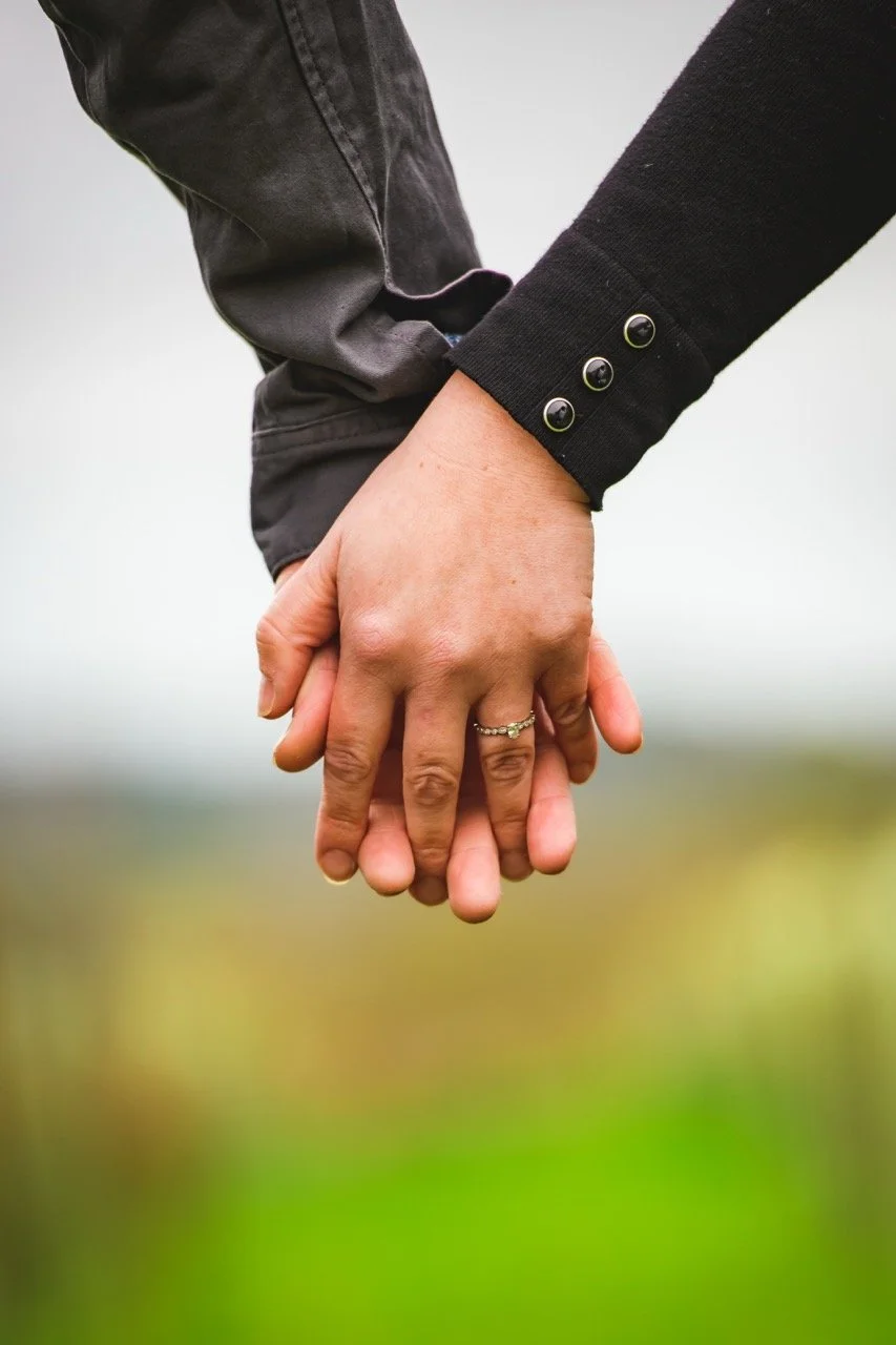 Close-up of a couple holding hands outdoors, with the woman wearing a ring and black sweater and the man wearing a dark shirt and jeans.