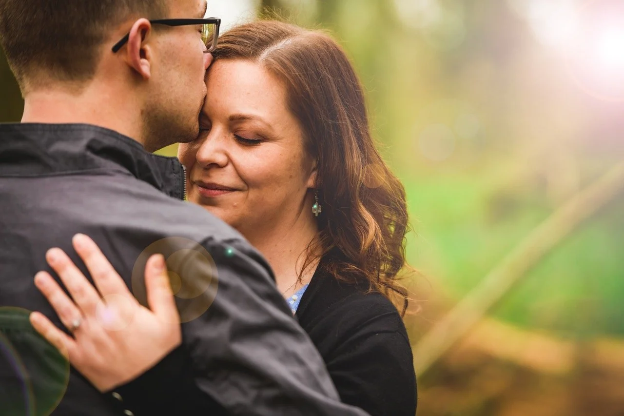 A man and woman embrace outdoors, with the man gently kissing the woman's forehead, both smiling softly with eyes closed.