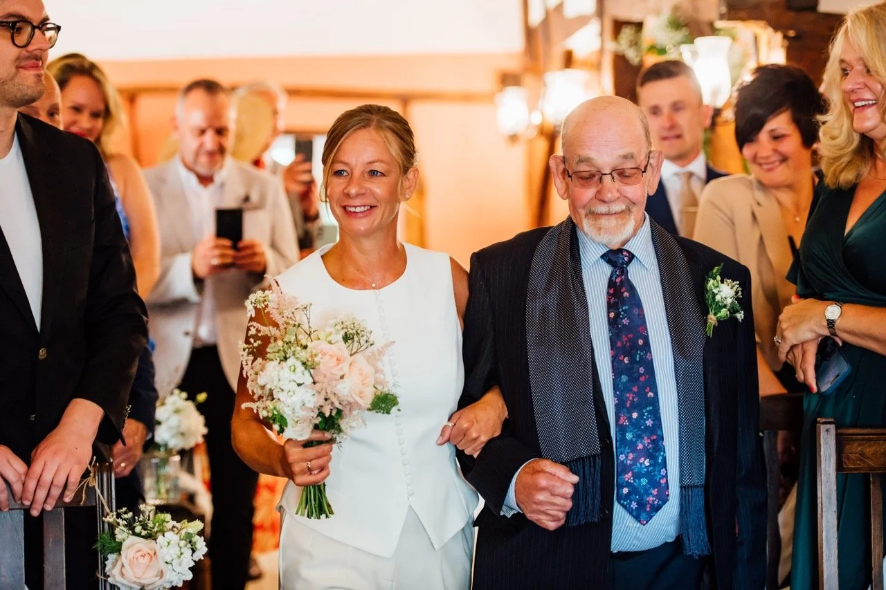 A bride walking down the aisle with an elderly man in a suit, holding a bouquet of flowers, surrounded by happy wedding guests in an indoor setting.