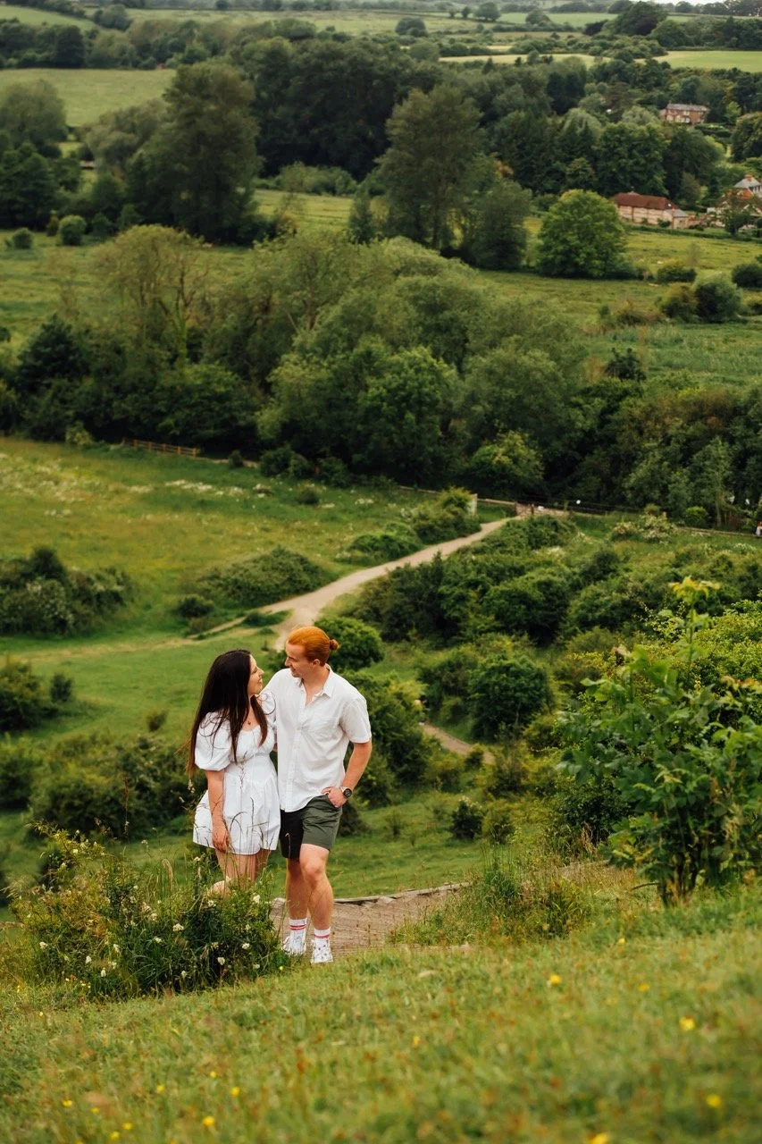 A young couple walking and talking on a trail in a lush green countryside with rolling hills, trees, and distant houses.