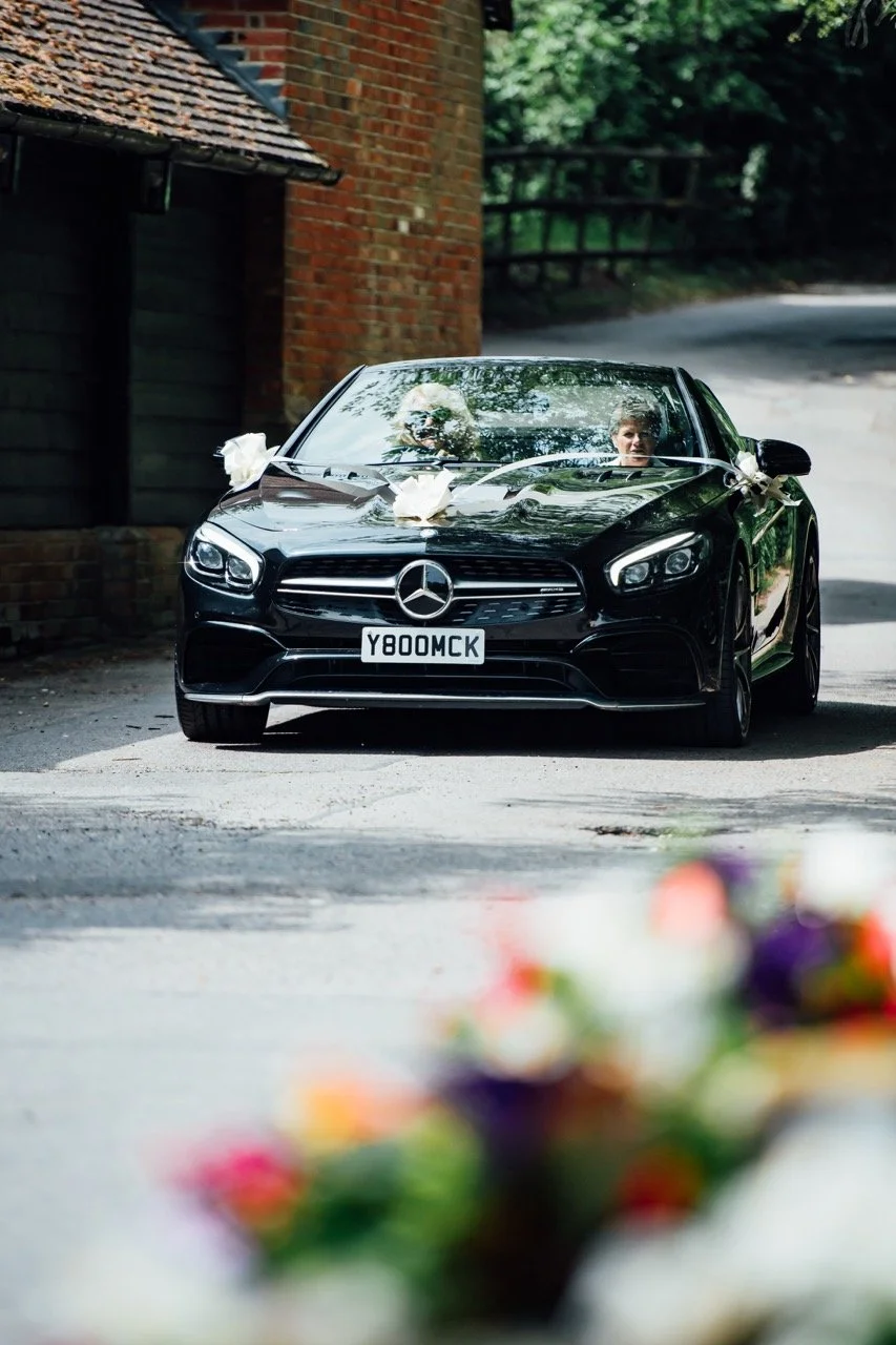 Black Mercedes-Benz car decorated with white ribbons and bows, parked on a narrow road near a brick building, with out-of-focus flowers in the foreground.