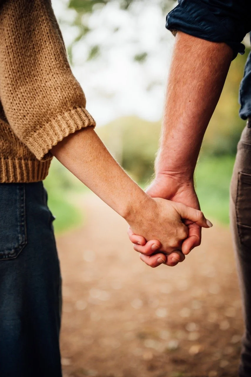 Close-up of a couple holding hands outdoors on a walking path surrounded by trees.