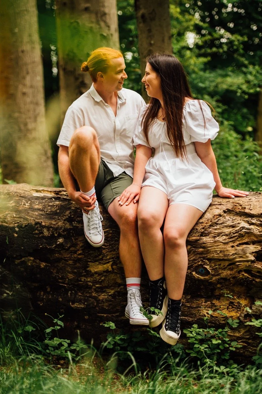 Two women sitting closely on a fallen tree in a lush green forest, smiling and looking at each other.