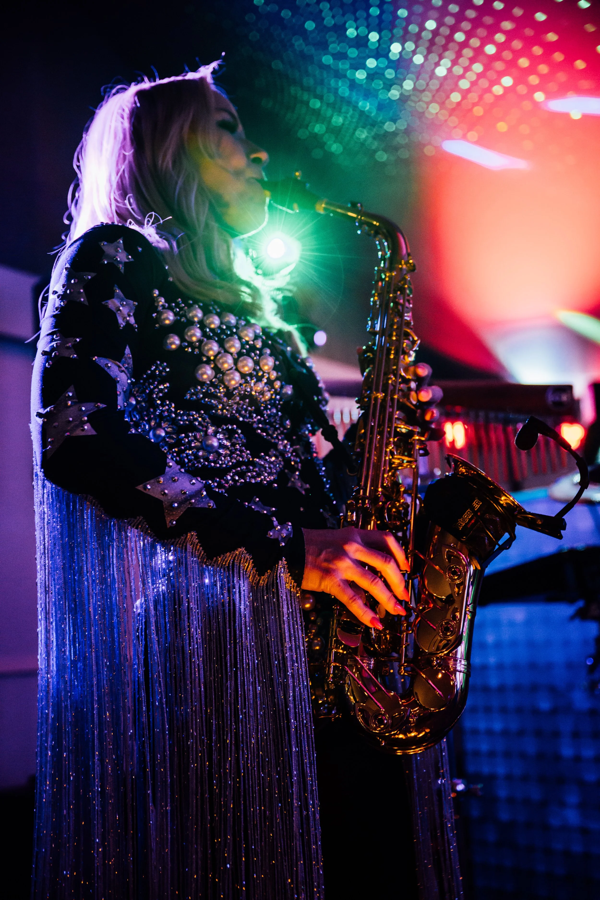A woman playing a saxophone on stage, illuminated by colorful lights, wearing a glittery, embellished dress with pearls and star patterns.
