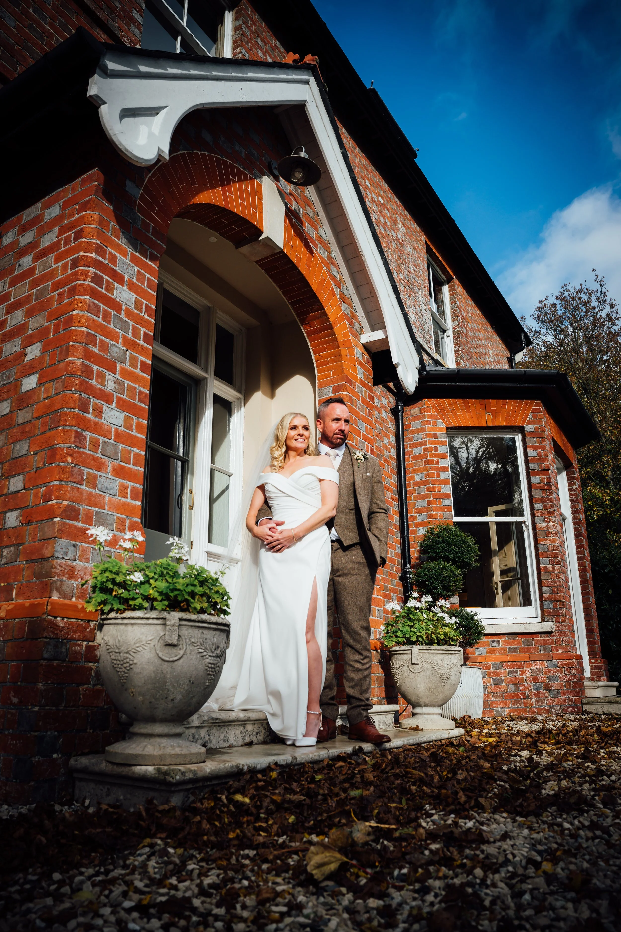 A bride and groom standing on the steps of a brick house, with the bride in a white dress and the groom in a brown suit, under a white decorative porch roof, with potted plants nearby.