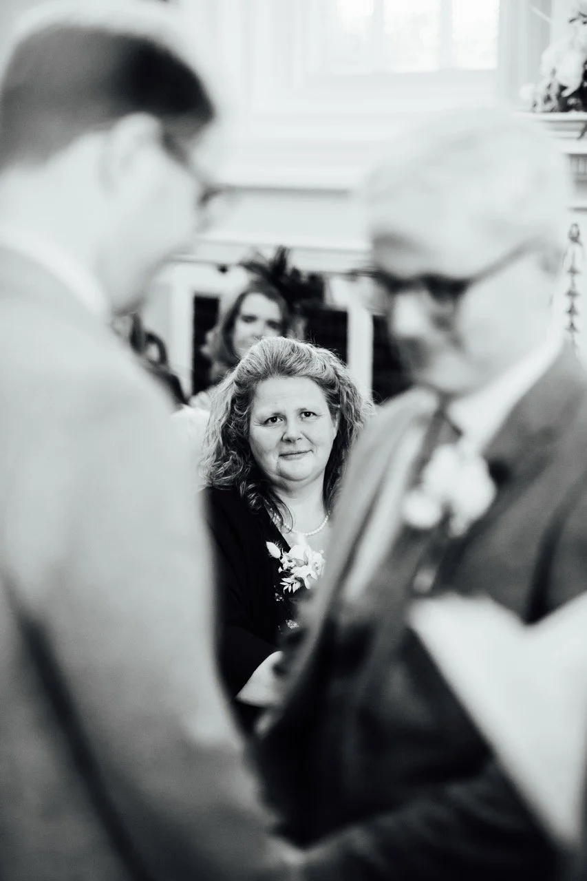 Black and white photograph of three people in a social setting. One woman with curly hair and a floral boutonniere looks directly at the camera, while two other individuals are blurred in the foreground and engaged in conversation.