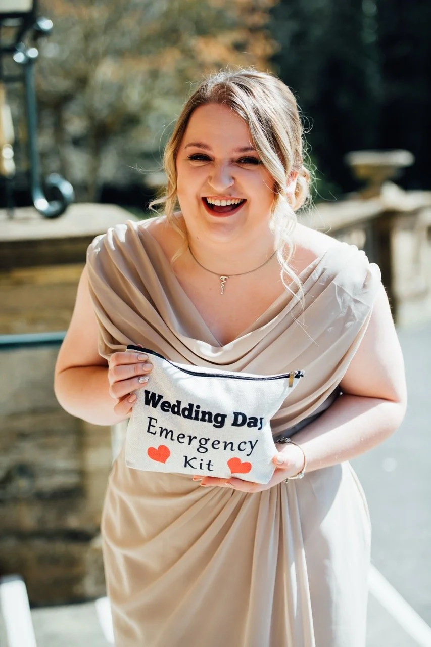 A woman in a beige dress holding a small pouch labeled 'Wedding Day Emergency Kit' with hearts, outdoors with trees and a stone railing in the background.
