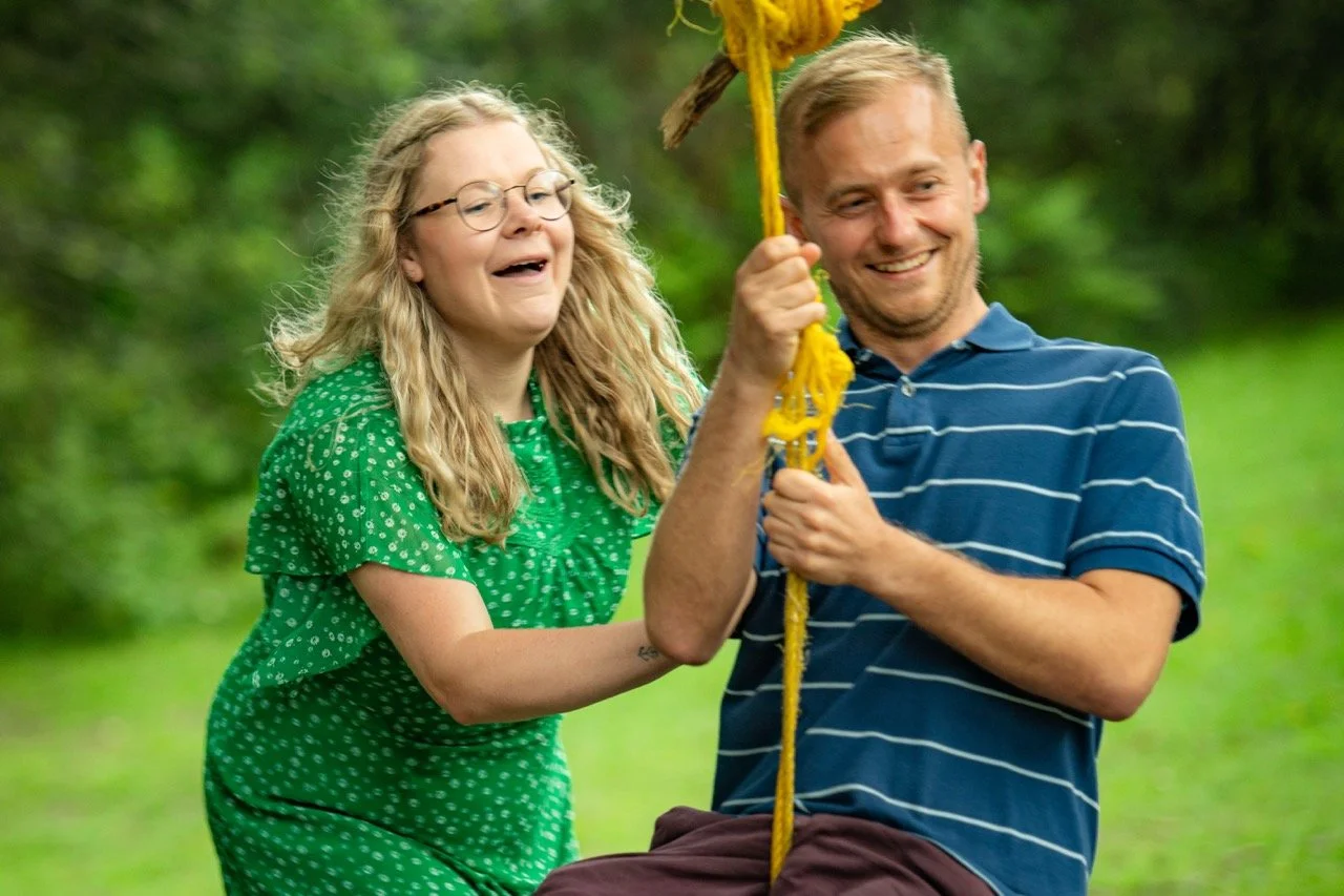 A woman in a green dress and a man in a blue striped shirt swinging together outdoors on a yellow rope swing amid lush green trees, both smiling and enjoying the moment.