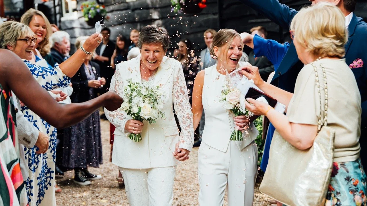 Two women in white outfits holding flowers walk hand-in-hand as they are celebrated with confetti at a wedding or celebration, surrounded by smiling guests.