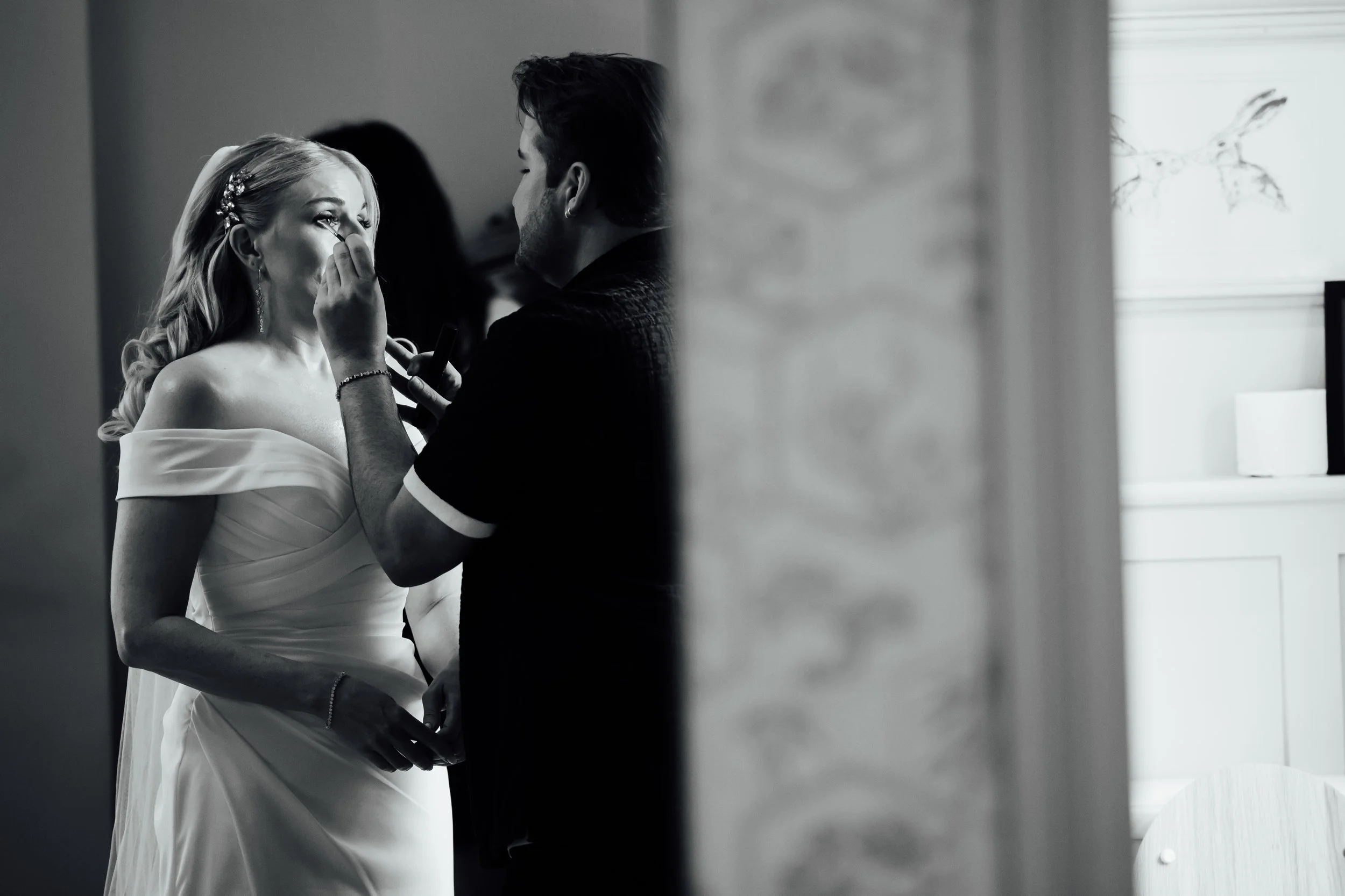 A bride getting her makeup done by a makeup artist in a room with natural light.