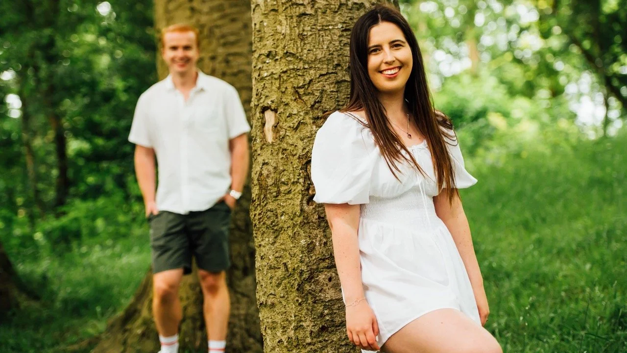 A young woman with long dark hair smiling, leaning against a tree in a forest, with a young man with red hair standing behind her, both dressed in casual white tops and dark shorts.