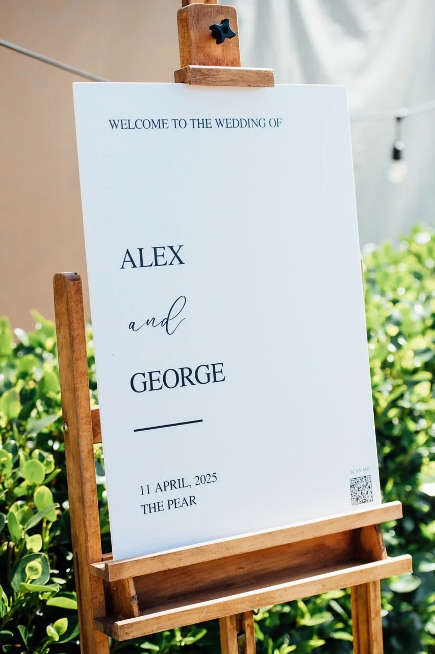 Wedding welcome sign with info about bride and groom, Alex and George, date April 11, 2025, at The Pear venue, on a wooden easel in a garden setting.