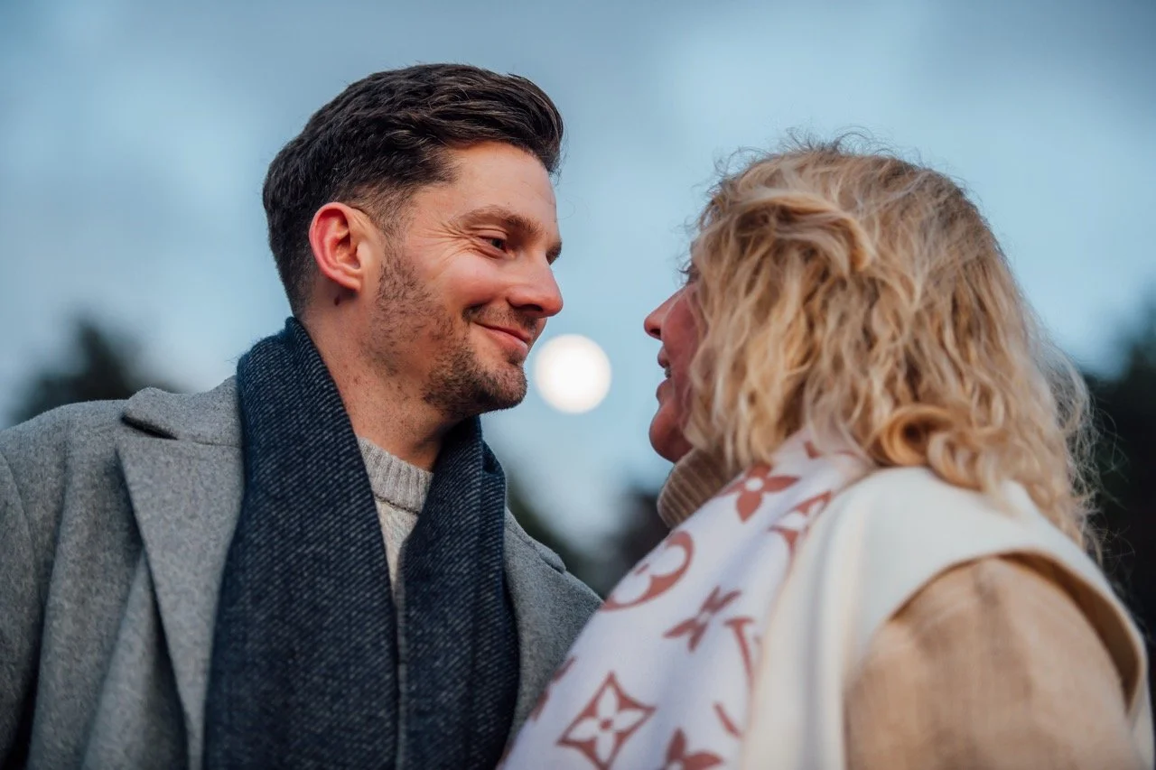 A young man and an older woman face each other outdoors, smiling and close to one another at dusk.