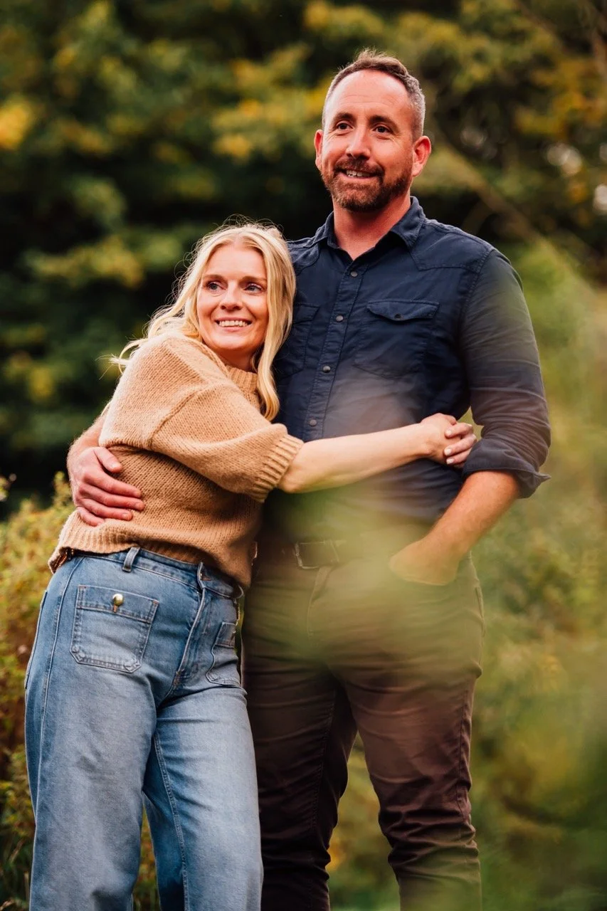 A smiling woman with blonde hair wearing a beige sweater hugging a man with short hair and a beard wearing a dark blue shirt, outdoors with greenery in the background.