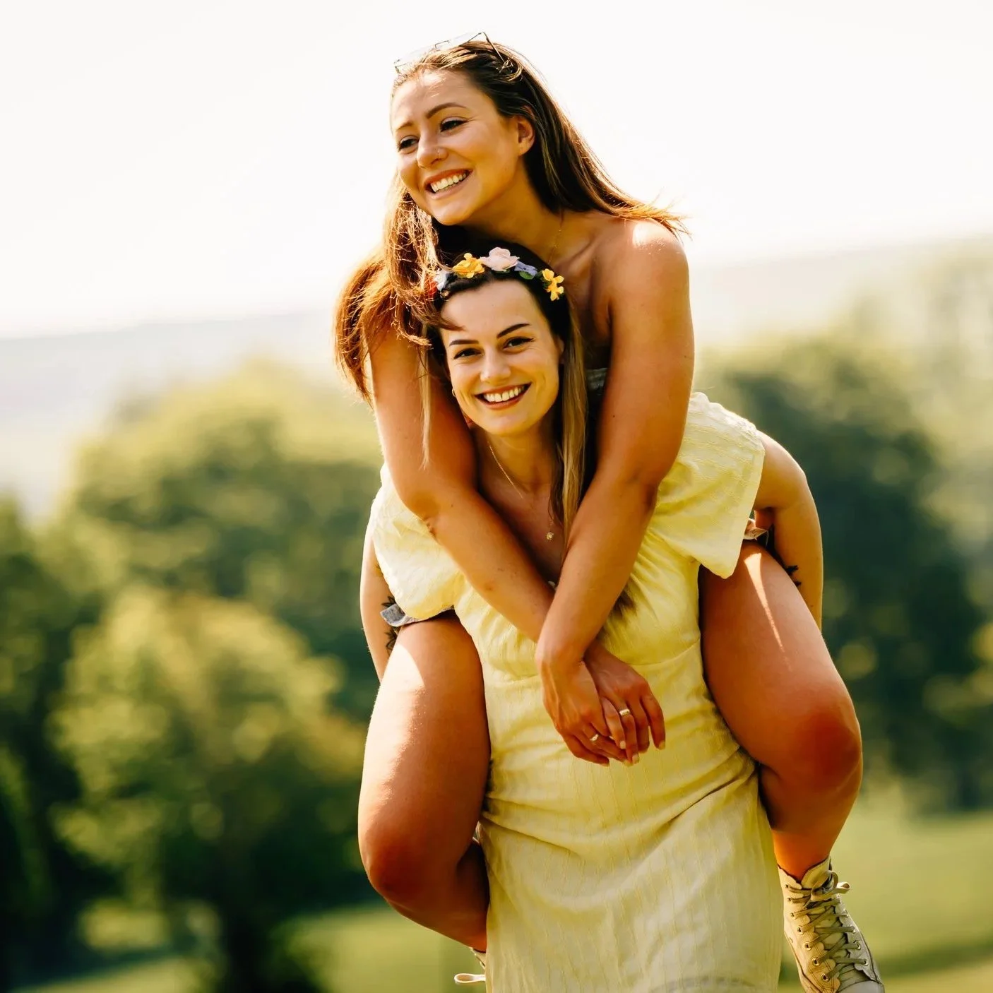 Two women outdoors, one giving the other a piggyback ride, both smiling and enjoying sunny weather with greenery in the background.