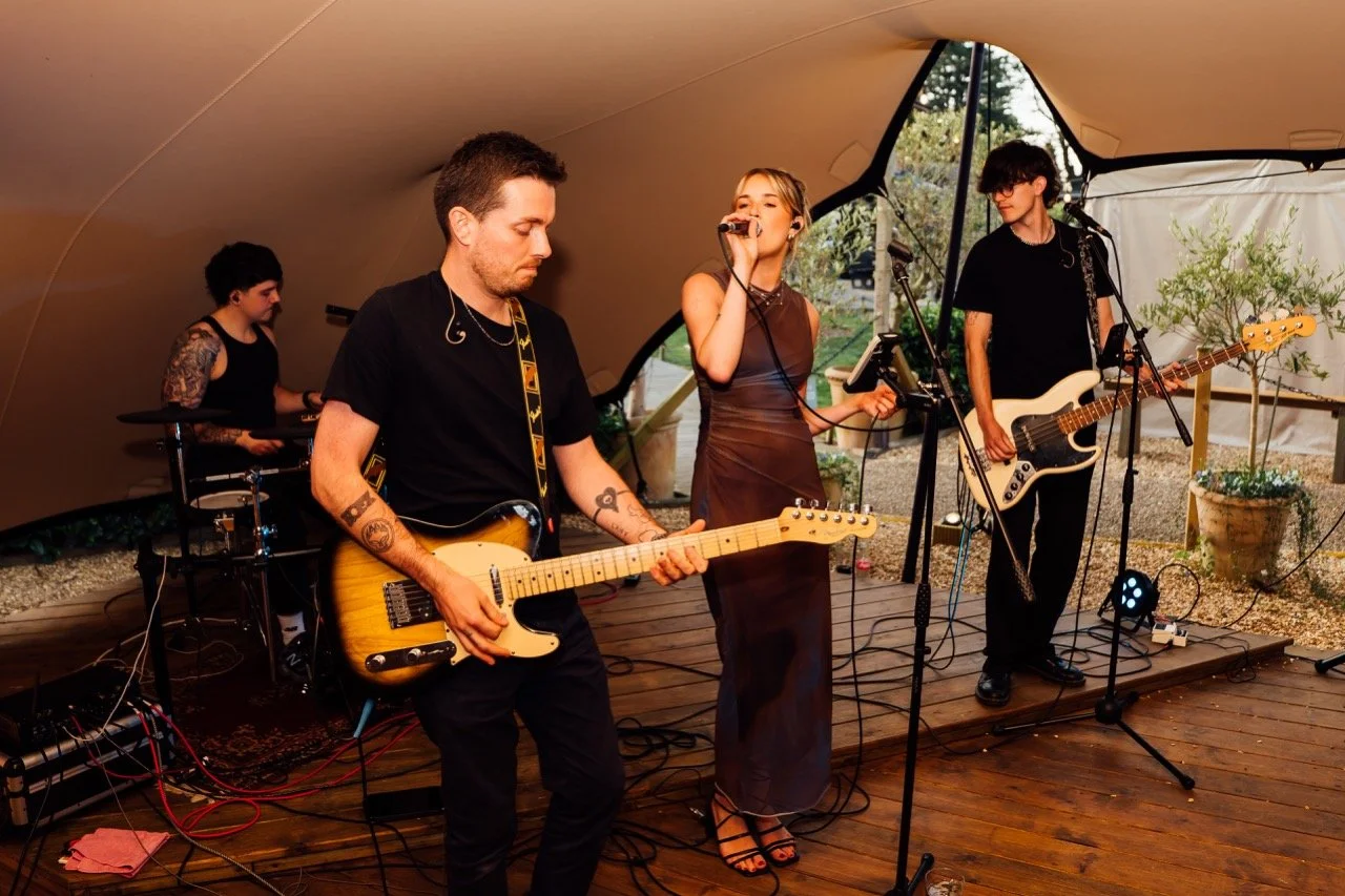 A band of four people performing under a canopy. The singer, a woman, is holding a microphone and singing. Two guitarists are playing electric guitars, and a drummer is playing drums in the background.