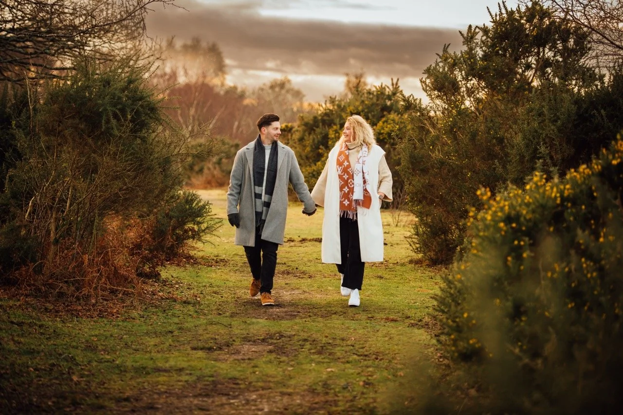 A young man and woman walking hand in hand on a nature trail surrounded by bushes and trees during late afternoon or early evening in fall or winter, dressed warmly with coats and scarves.