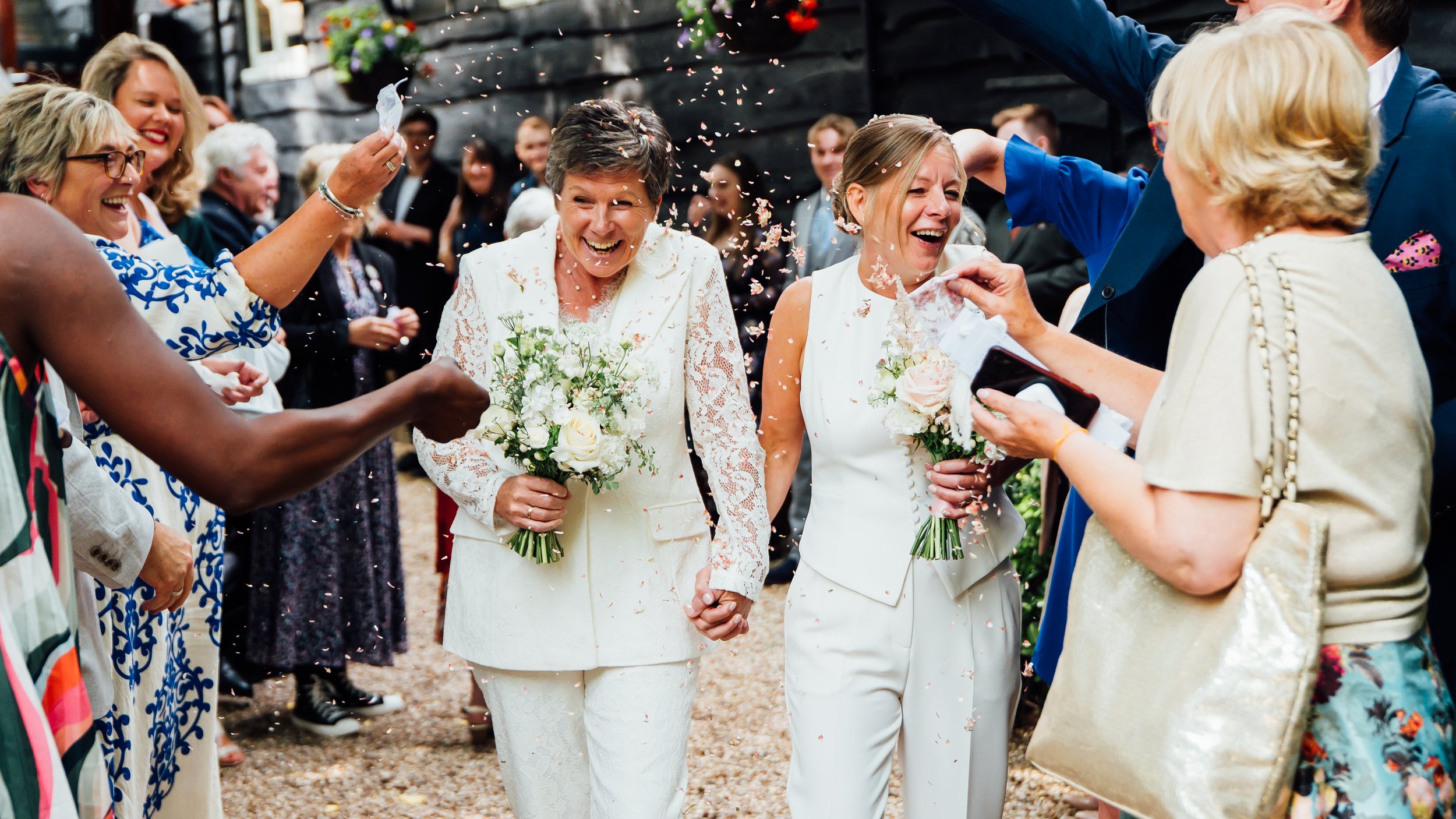 Two women in white wedding dresses holding bouquets of white flowers, smiling, walking hand in hand among a crowd of people celebrating and throwing confetti at an outdoor wedding reception.