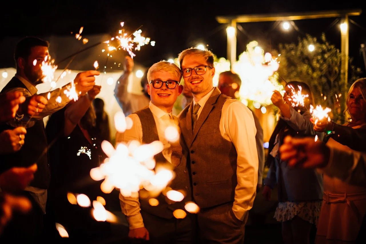 Two men dressed in formal attire smiling at a nighttime celebration surrounded by people holding sparklers.
