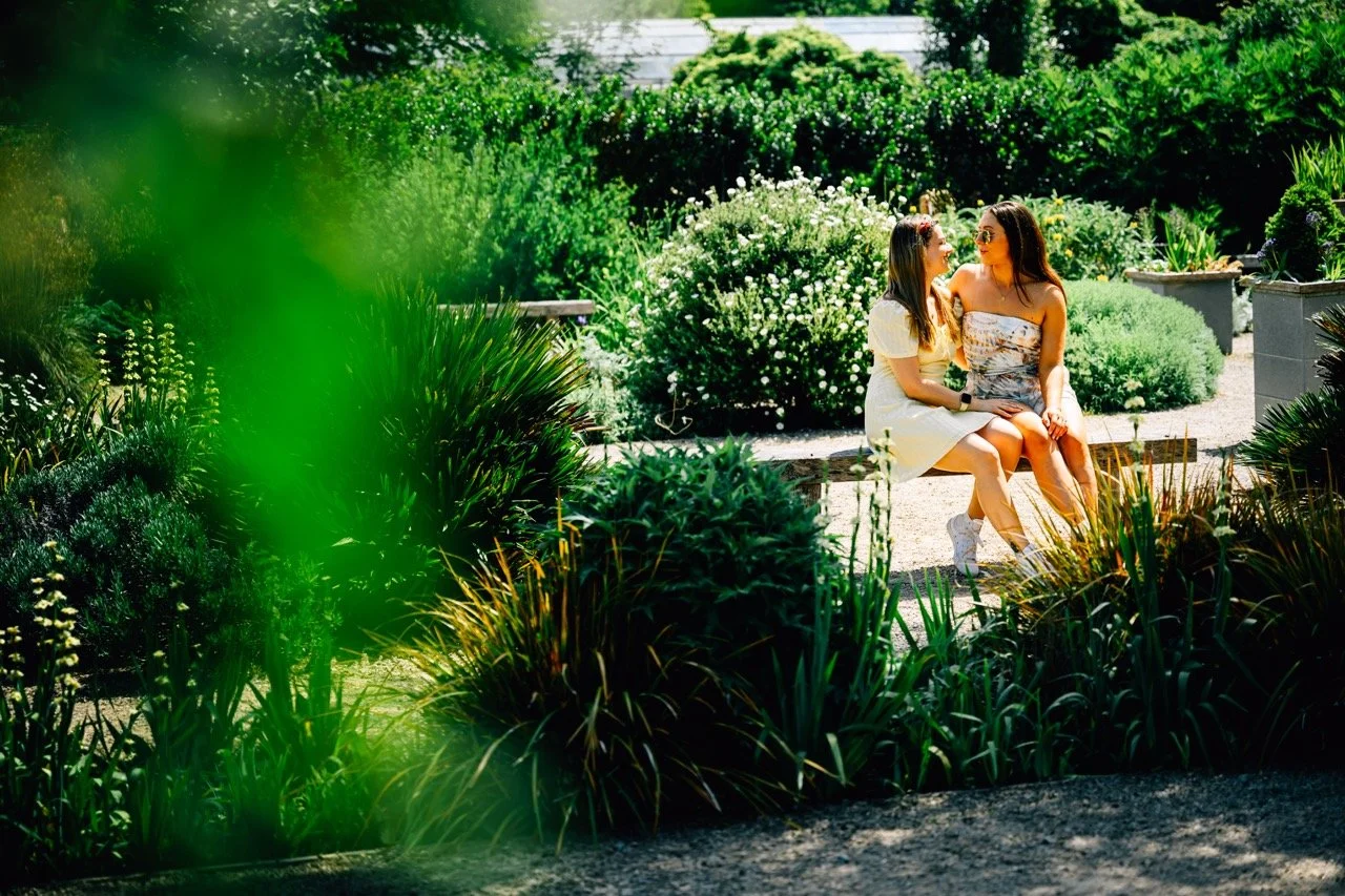 Two women sitting on a bench in a lush garden, engaging in conversation, surrounded by green plants and flowers.