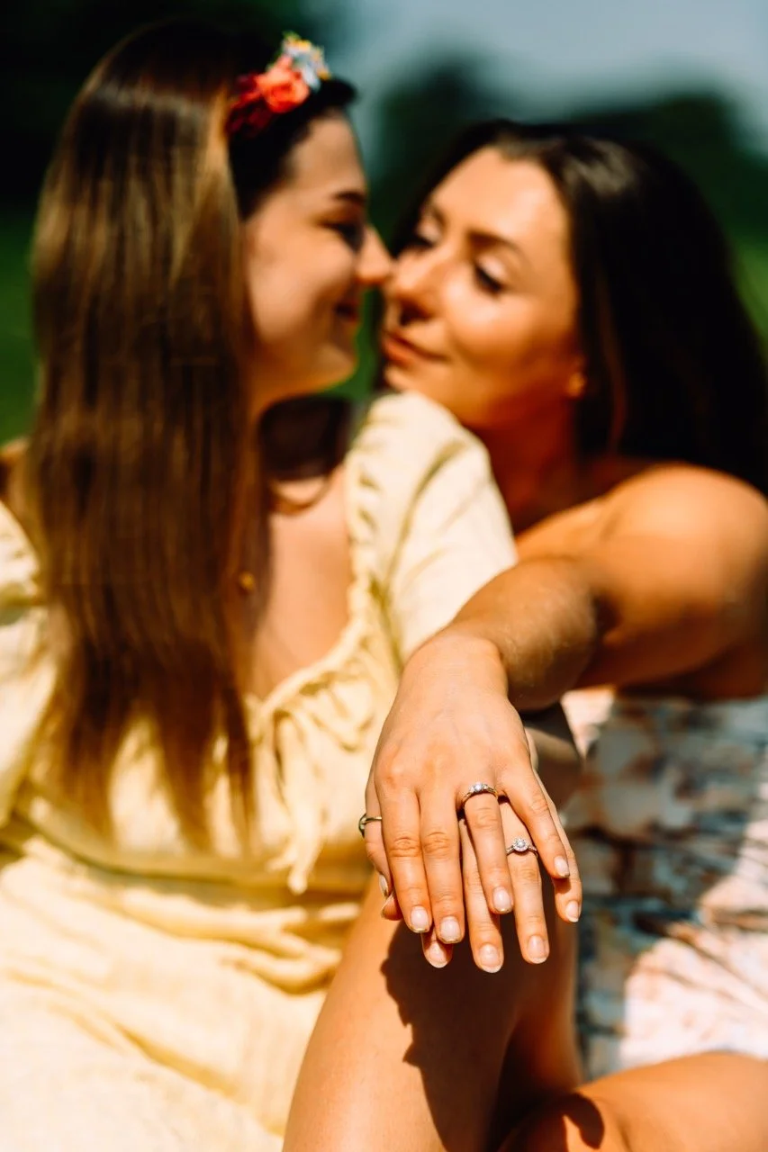 Two women sitting close together, touching foreheads and smiling warmly, with one woman showing off her wedding ring. The background is blurred with greenery.