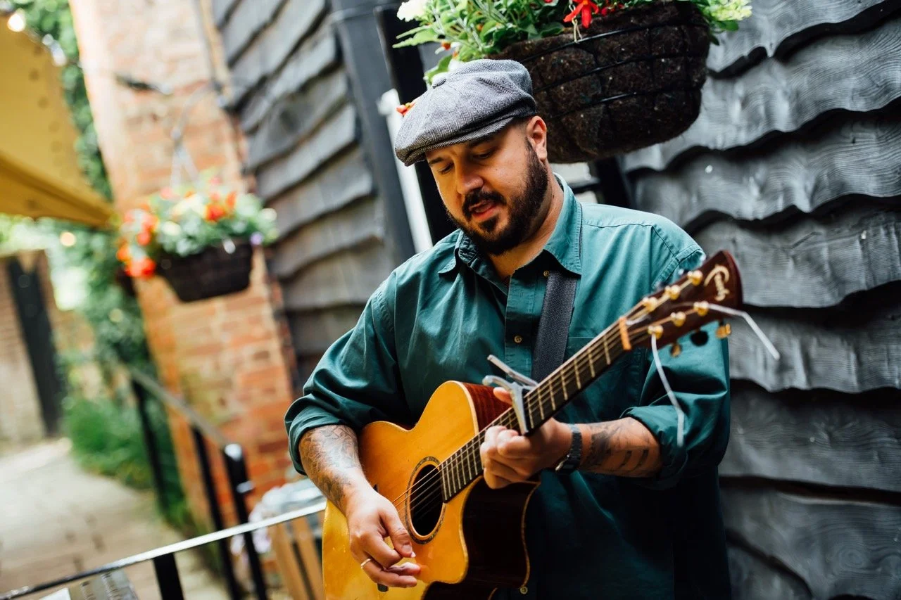 A man playing an acoustic guitar outdoors next to a black wood wall, wearing a gray cap and green shirt, with floral hanging baskets visible in the background.