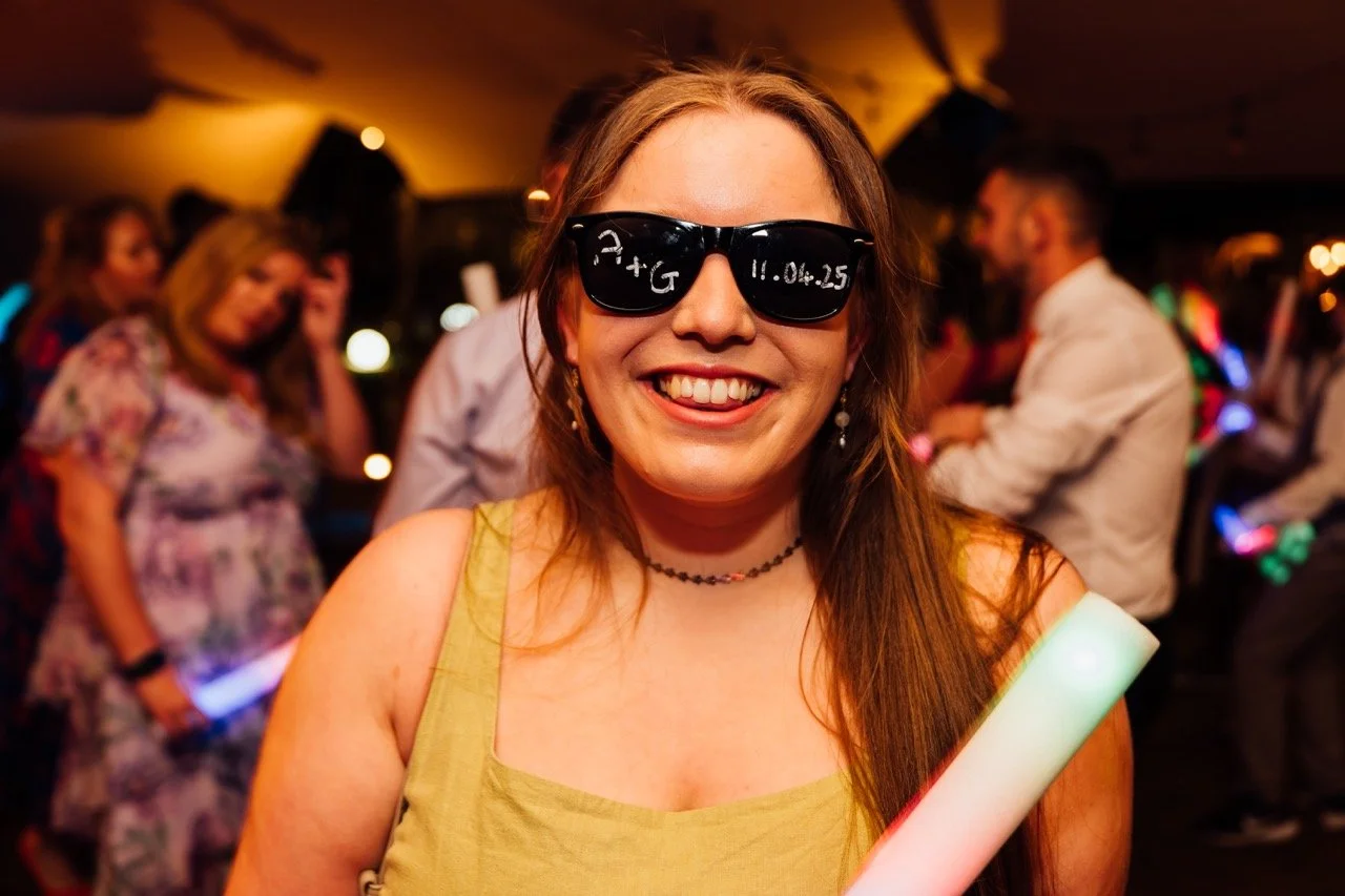 A smiling woman in sunglasses at a party or celebration, holding a glow stick with other people dancing and socializing in the background.