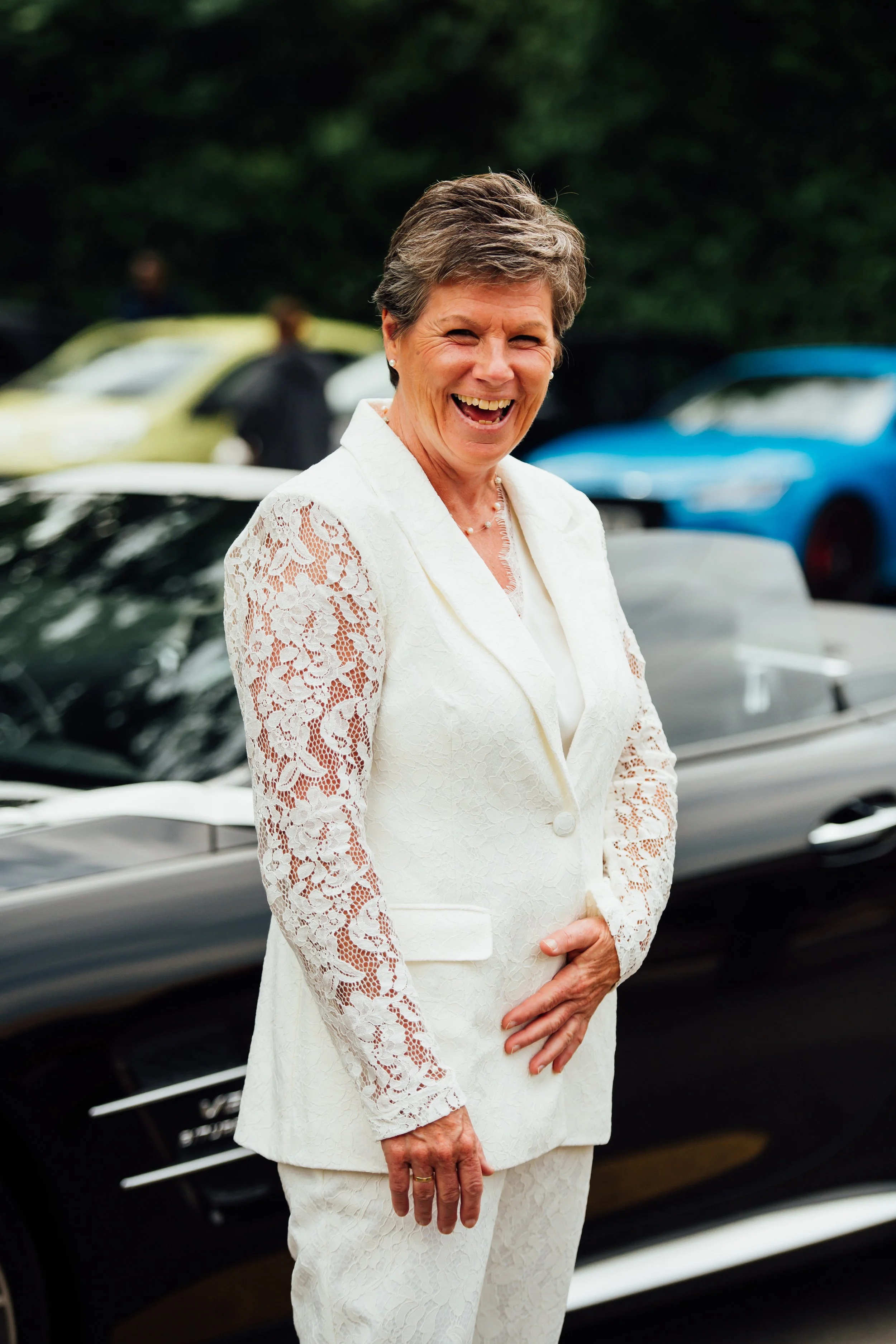 A smiling older woman with short gray hair wearing a white lace blazer and matching pants, standing outdoors in front of parked cars and trees.
