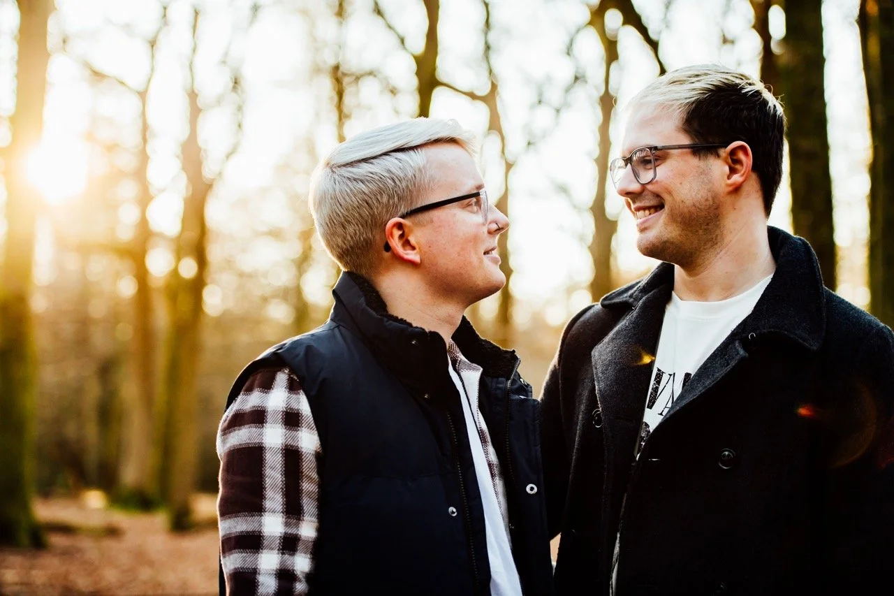 Two young men with glasses face each other smiling in a wooded park during sunset.