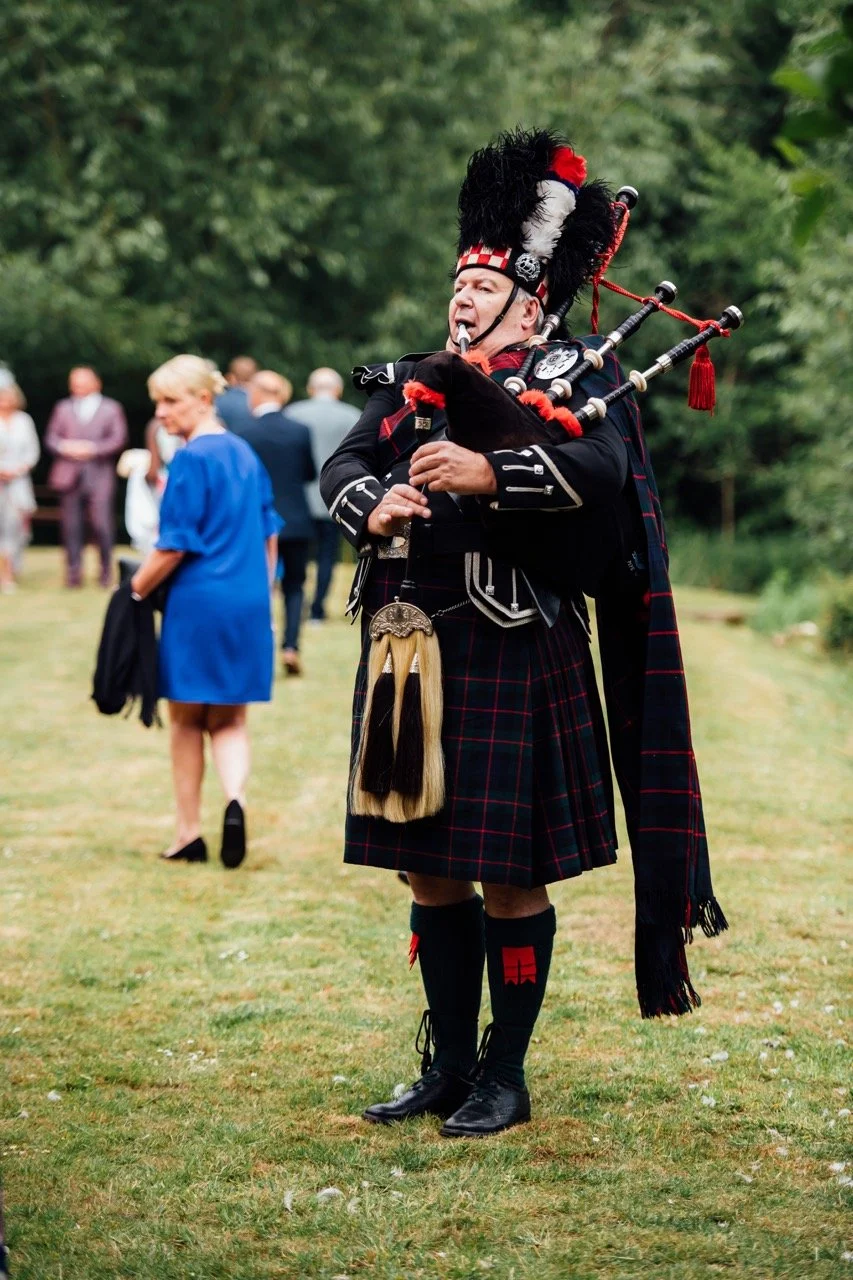 Man dressed in traditional Scottish attire playing bagpipes at an outdoor event, with people in formal clothing in the background.