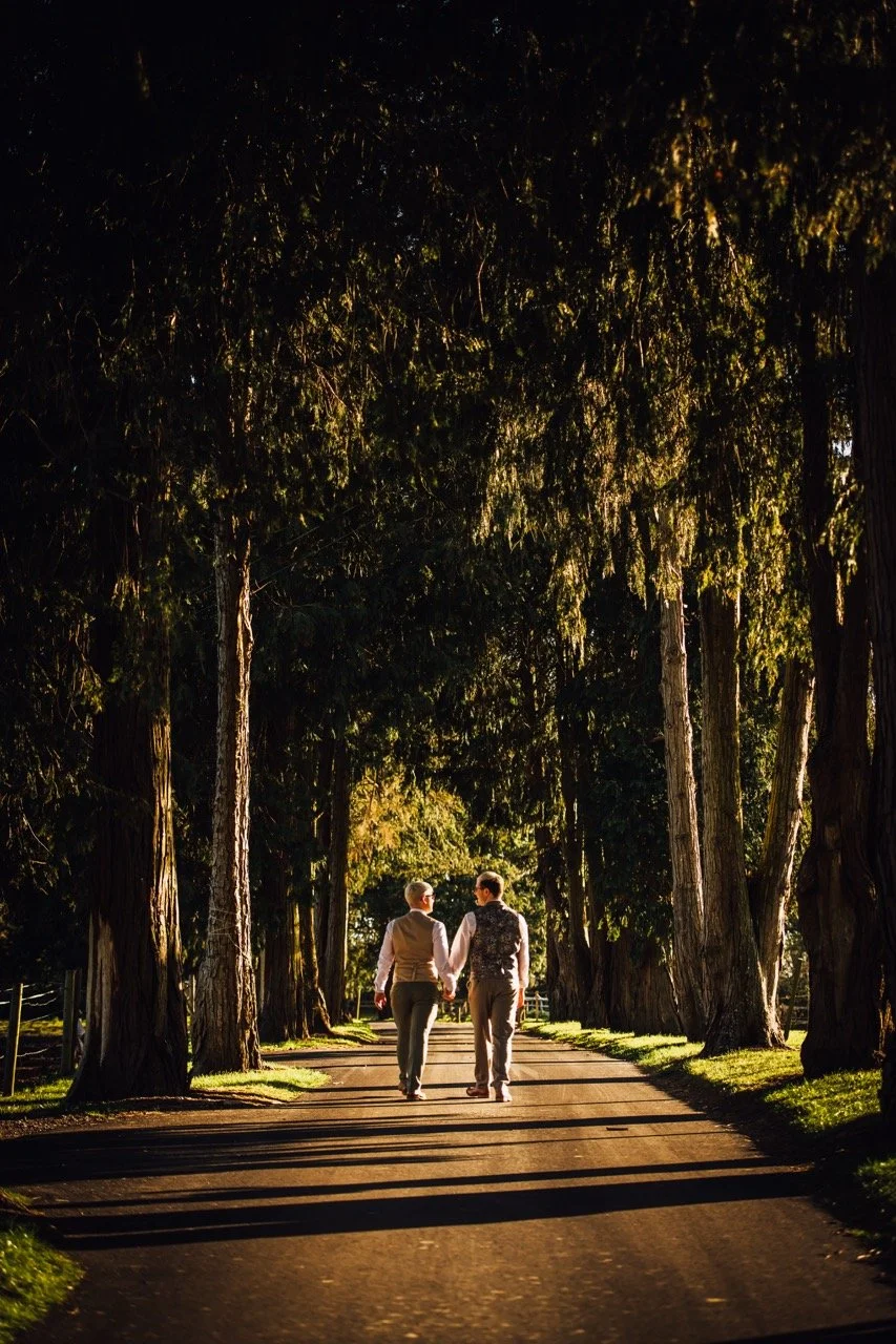A couple holding hands walking down a wooded path during sunset, surrounded by tall trees.