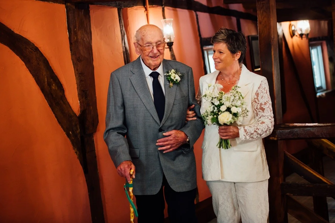 An elderly man and a middle-aged woman, possibly a bride, are standing together indoors. The man is dressed in a gray checkered suit with a white shirt, black tie, and glasses, and is holding a colorful walking cane. The woman is wearing a white lace