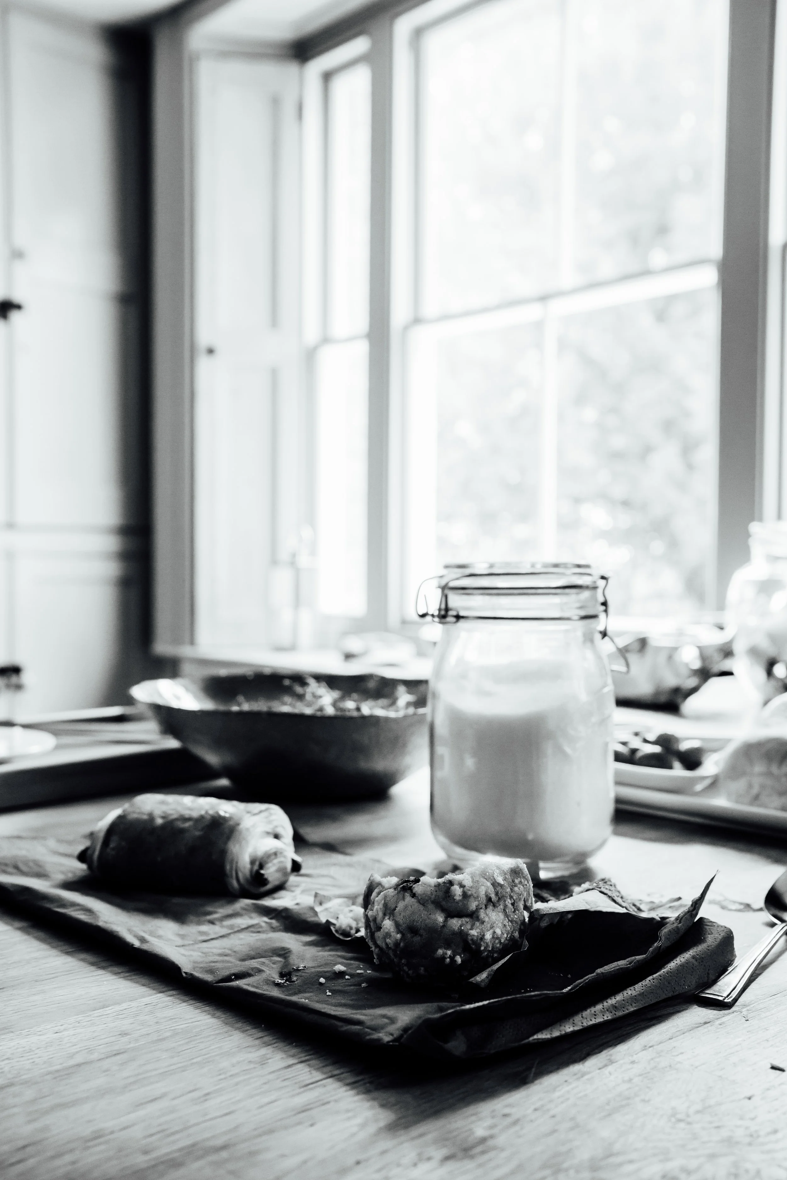 A kitchen table with a rolled pastry, a muffin, and a jar of flour in front of a window.