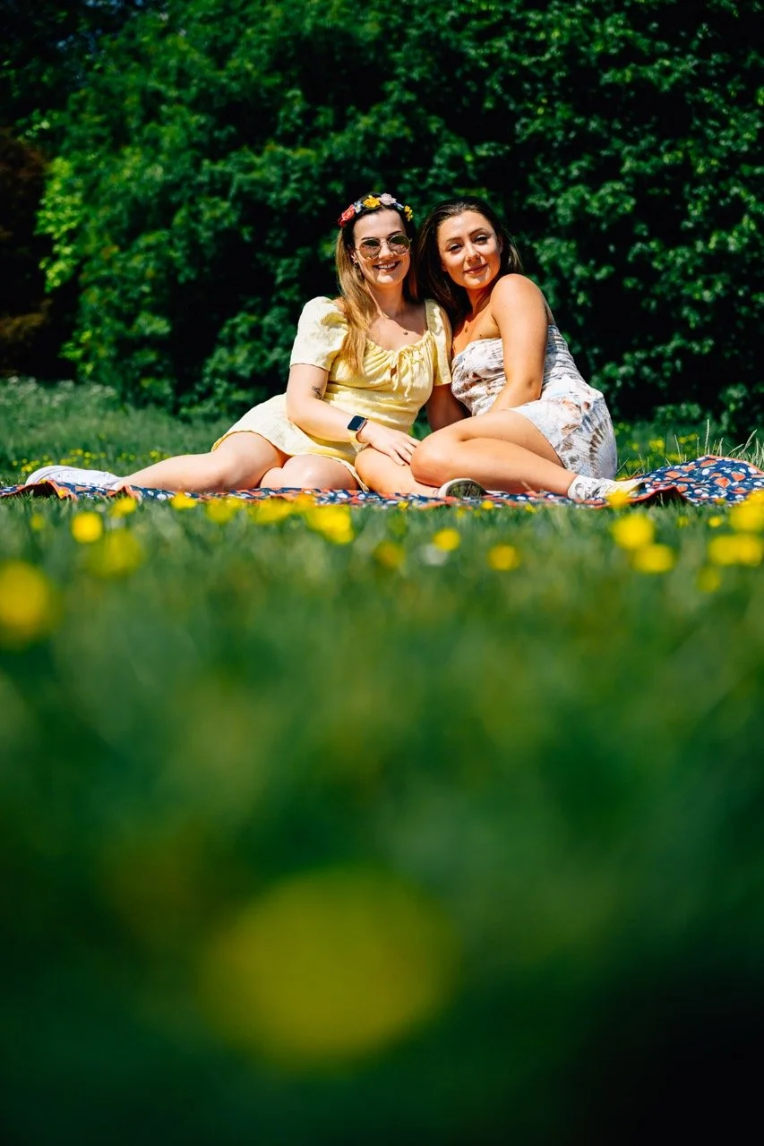Two women sitting on a blanket outdoors on the grass, surrounded by trees, enjoying a sunny day. One woman is wearing a yellow dress with a flower headband and sunglasses, while the other is wearing a strapless dress. They are smiling and appear happ